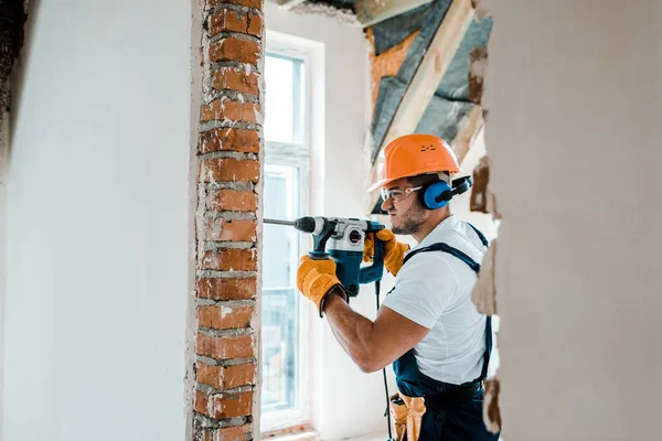 Enfoque selectivo del trabajador en guantes uniformes y amarillos con taladro de martillo en la pared de ladrillo - foto de stock