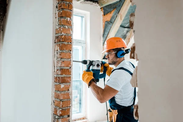 Messa a fuoco selettiva del lavoratore in uniforme e guanti gialli con trapano a martelli sulla parete di mattoni — Foto stock