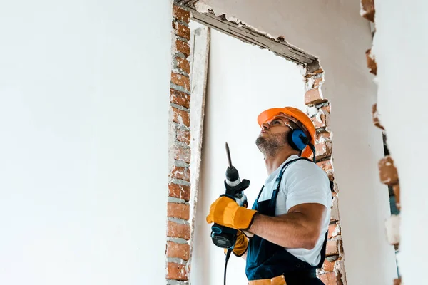 Obrero guapo sosteniendo taladro de martillo y mirando a la pared - foto de stock