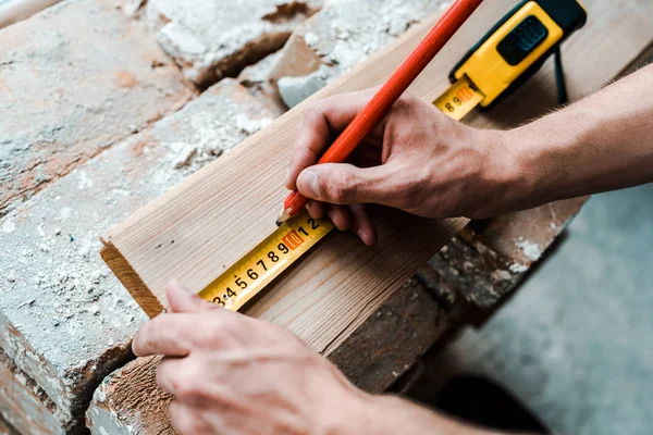 Vista recortada del reparador sosteniendo lápiz mientras mide tablón de madera - foto de stock