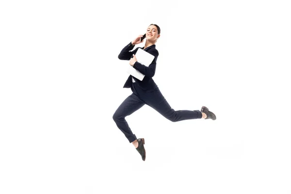 Positive businesswoman talking on smartphone and holding laptop while levitating and looking at camera isolated on white — Stock Photo