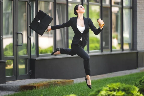 Счастливая бизнесвумен, левитирующая, держа в руках чемодан и кофе, чтобы пойти — стоковое фото