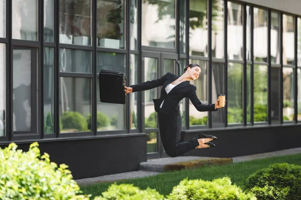 Fröhliche Geschäftsfrau tanzt in der Nähe von Bürogebäuden und hält Aktentasche und Coffee to go in der Hand — Stockfoto