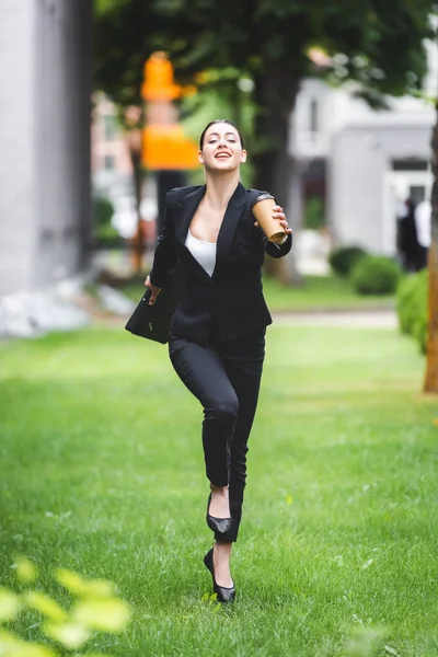 Alegre, atractiva mujer de negocios corriendo en el césped mientras sostiene el café para ir - foto de stock