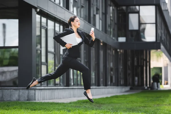 Attractive businesswoman levitating while using smartphone and holding laptop — Stock Photo