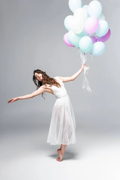 Young attractive ballerina in white dress dancing with festive balloons on grey background — Stock Photo