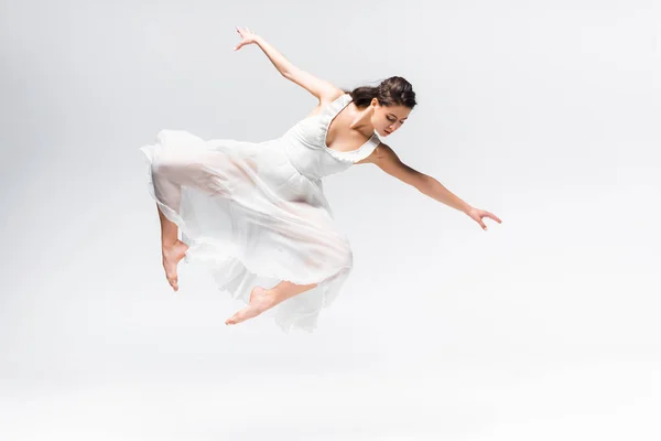 Красива балерина в білій сукні, стрибає в танці на сірому фоні — стокове фото