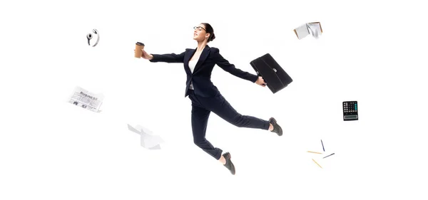 Plano panorámico de la mujer de negocios sosteniendo café para ir y maletín mientras levita auriculares rodeados, periódico, calculadora y papelería aislado en blanco - foto de stock