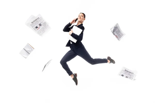 Sonriente mujer de negocios hablando en un smartphone y sosteniendo el portátil mientras levita rodeado de periódicos volando aislados en blanco - foto de stock