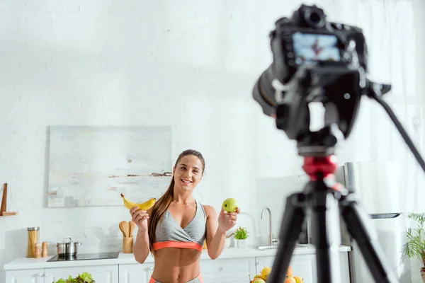 Enfoque selectivo de la mujer feliz sosteniendo manzana y plátano cerca de la cámara digital - foto de stock