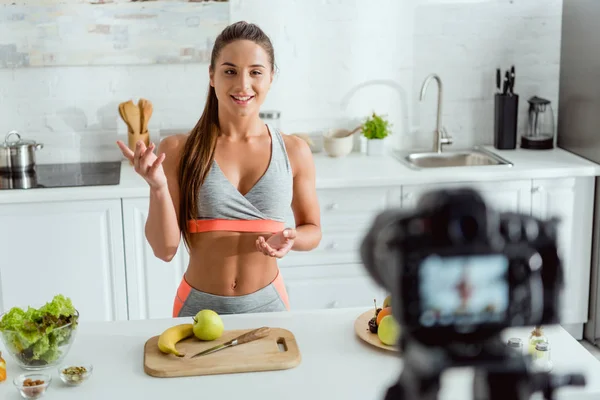 Selective focus of happy video blogger gesturing near fruits and digital camera — Stock Photo