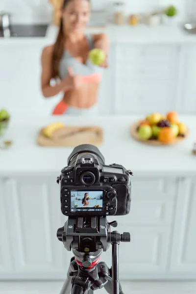 Selective focus of digital camera with happy woman holding apple and showing thumb up on screen — Stock Photo