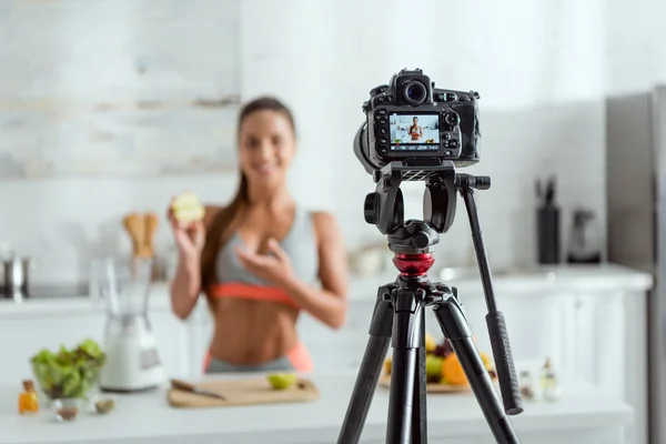 Selective focus of digital camera with happy woman holding half of apple and gesturing on screen — Stock Photo