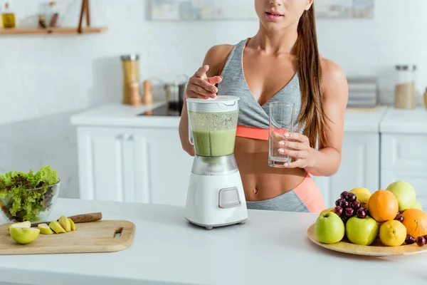 Cropped view of sportswoman standing near blender with smoothie and fruits — Stock Photo