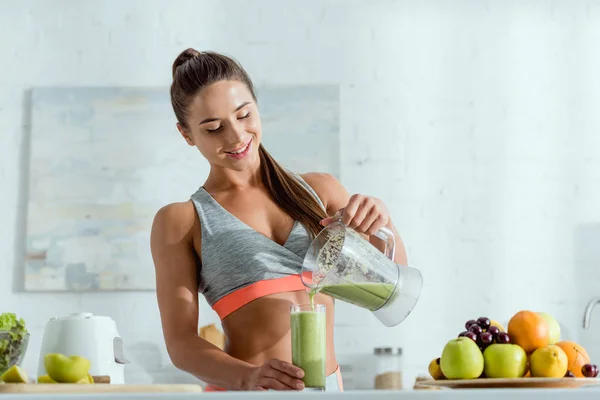 Cheerful girl with sportswear pouring smoothie in glass from blender — Stock Photo