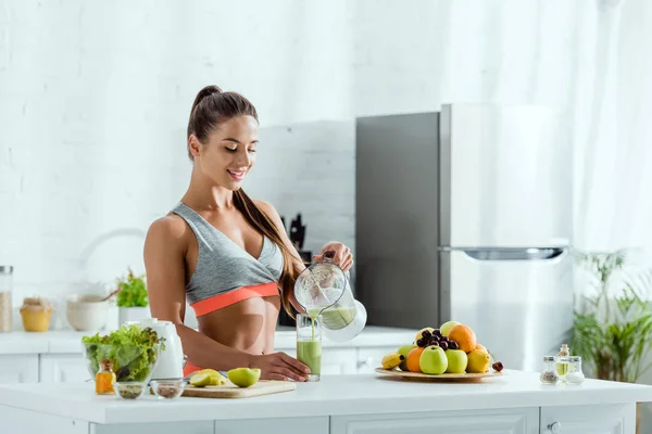 Femme heureuse avec des vêtements de sport verser smoothie dans le verre près des fruits — Photo de stock