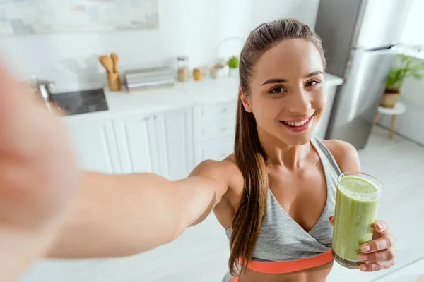 Selective focus of happy girl holding green smoothie and looking at camera — Stock Photo