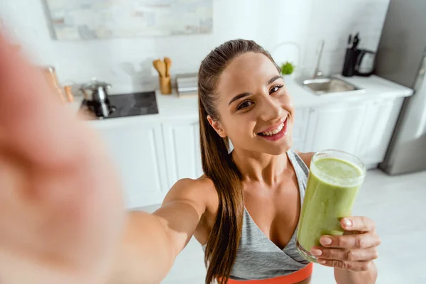 Selective focus of positive girl holding green smoothie and looking at camera — Stock Photo