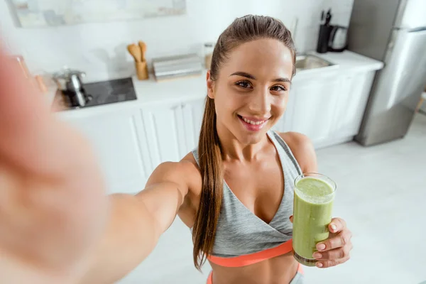 Selective focus of smiling girl holding glass with green smoothie and looking at camera — Stock Photo