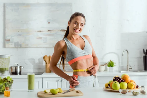 Mujer feliz y deportiva sosteniendo cinta métrica mientras mide la cintura cerca de las frutas - foto de stock