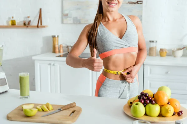 Vista recortada de la mujer joven que mide la cintura cerca de los frutos y muestra el pulgar hacia arriba - foto de stock
