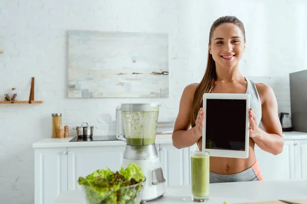 Mujer feliz sosteniendo tableta digital con pantalla en blanco cerca de licuadora y vidrio con batido - foto de stock