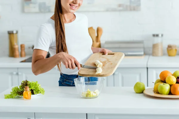 Cropped view of happy young woman putting banana in bowl with apples — Stock Photo