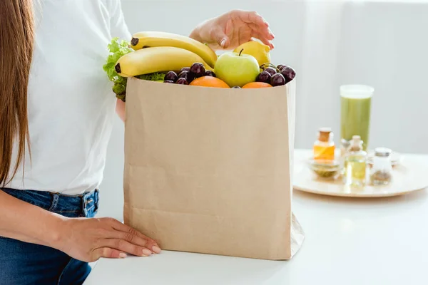 Vista recortada de la joven mujer de pie cerca de la bolsa de papel con frutas — Stock Photo