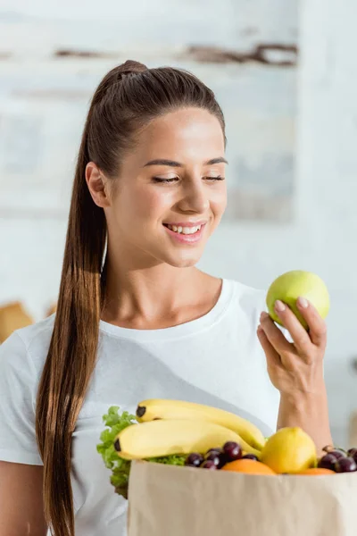 Jovem mulher positiva olhando para maçã verde perto de saco de papel com frutas — Fotografia de Stock