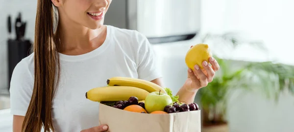Панорамный снимок положительной женщины, смотрящей на лимон возле бумажного пакета с фруктами — стоковое фото