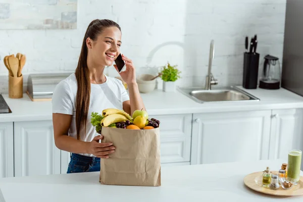 Mujer feliz hablando en el teléfono inteligente cerca de bolsa de papel con frutas - foto de stock