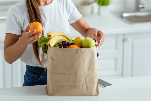 Cropped view of woman with orange near paper bag and smartphone — Stock Photo