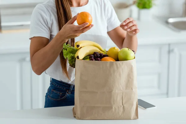 Cropped view of woman with orange near paper bag and smartphone with blank screen — Stock Photo