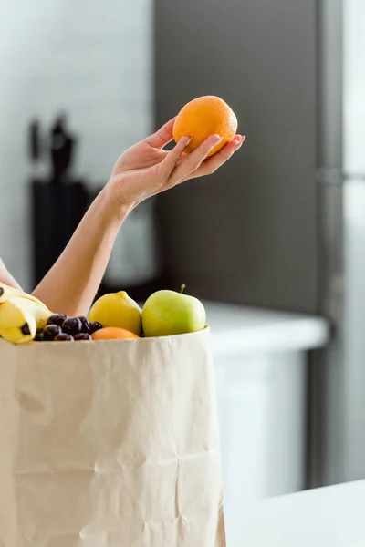 Vista recortada de la mujer sosteniendo naranja fresca y orgánica cerca de la bolsa de papel con frutas - foto de stock