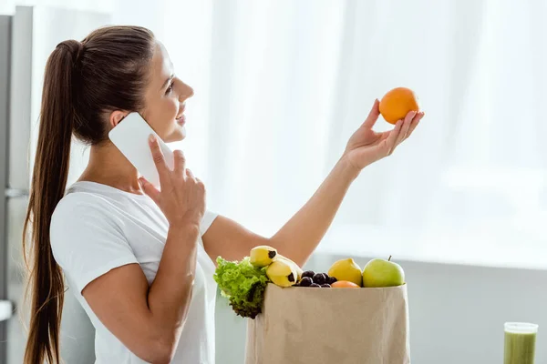 Donna felice che parla su smartphone vicino sacchetto di carta con generi alimentari — Foto stock