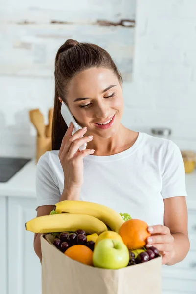 Joyeuse jeune femme parlant sur smartphone près du sac en papier avec épicerie — Photo de stock