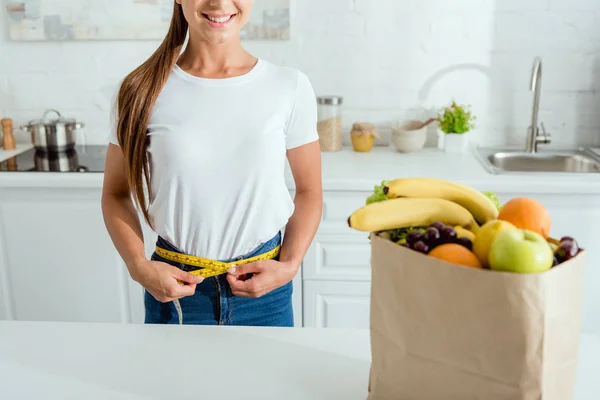 Vue recadrée d'une jeune femme joyeuse mesurant la taille près d'un sac en papier avec épicerie — Photo de stock