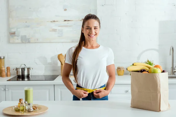 Cheerful young woman measuring waist near paper bag with groceries — Stock Photo