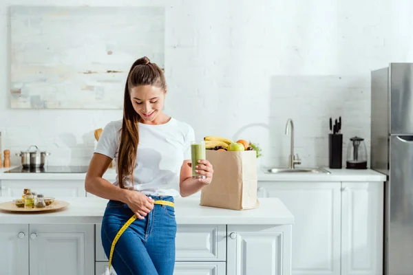 Happy young woman measuring waist and holding glass with smoothie near groceries — Stock Photo