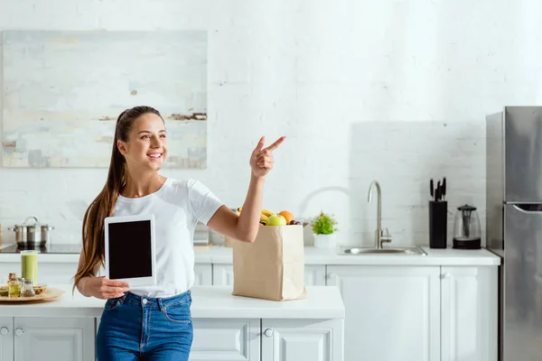 Chica alegre sosteniendo tableta digital con pantalla en blanco y apuntando con el dedo cerca de comestibles - foto de stock