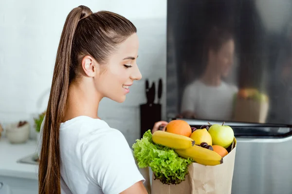 Selective focus of young happy woman opening fridge and holding groceries — Stock Photo
