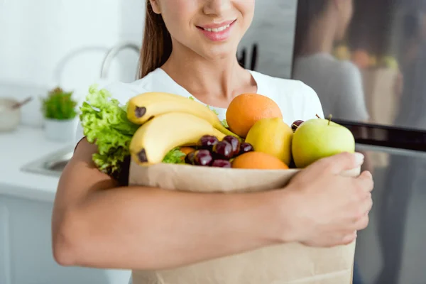 Cropped view of cheerful girl holding paper bag with groceries near fridge — Stock Photo