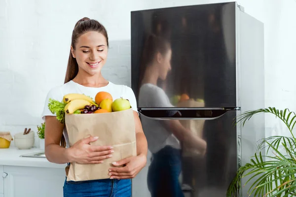 Веселая девушка держит бумажный пакет с продуктами возле холодильника — стоковое фото