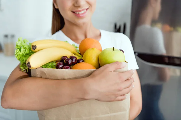 Vista recortada de niña alegre sosteniendo bolsa de papel con deliciosos comestibles — Stock Photo