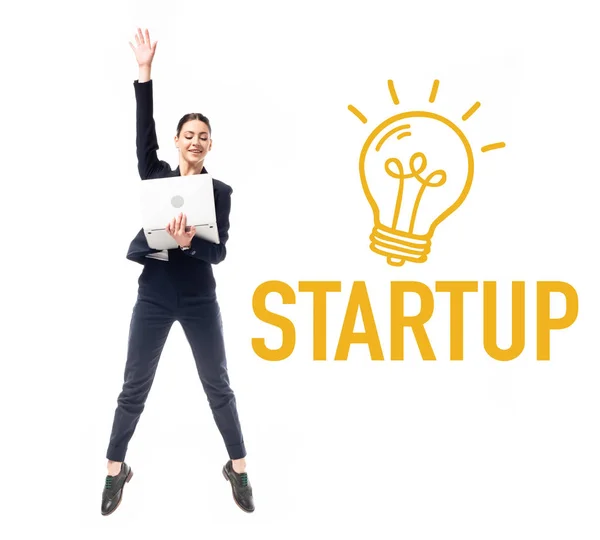 Smiling businesswoman using laptop while jumping near startup word and drawn electric bulb isolated on white — Stock Photo