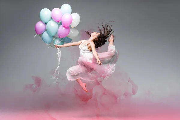 Bella ballerina che balla con palloncini festivi vicino a spruzzi di fumo rosa su sfondo grigio — Foto stock
