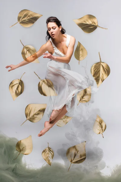 Beautiful ballerina in white dress dancing surrounded by falling leaves near grey smoke on grey background — Stock Photo