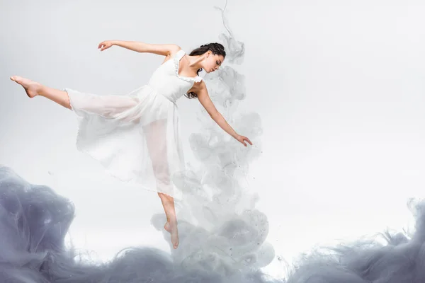 Young graceful ballerina in white dress dancing in grey smoke on grey background — Stock Photo