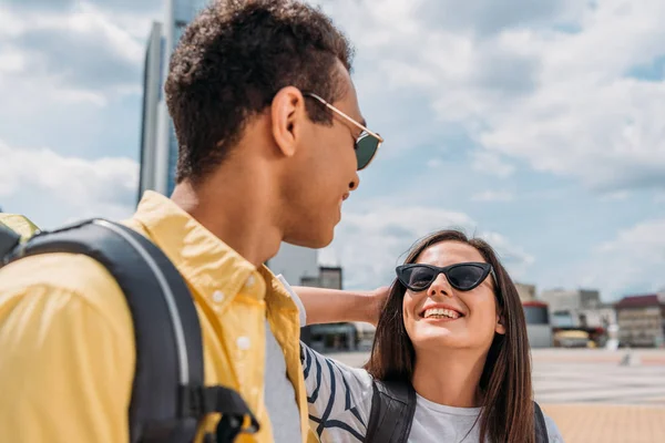 Mujer con gafas de sol sonriendo y mirando a un amigo bi-racial con mochila - foto de stock