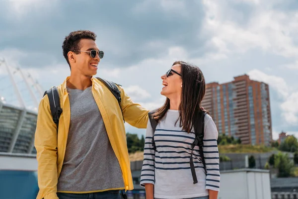 Cheerful interracial friends in sunglasses looking at each other and smiling with backpacks — Stock Photo